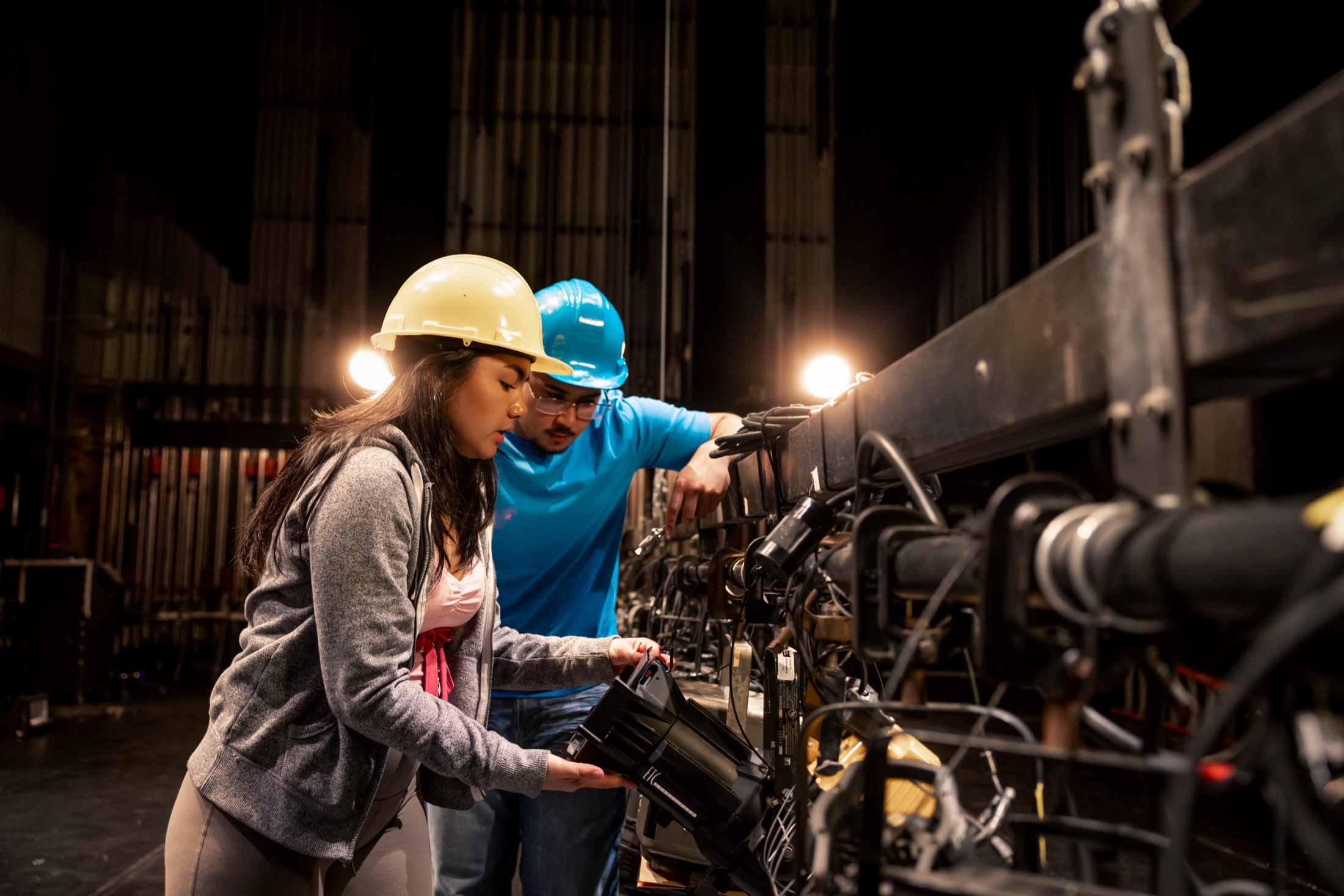 Two students in hard hats focusing a lighting instrument on a batten backstage