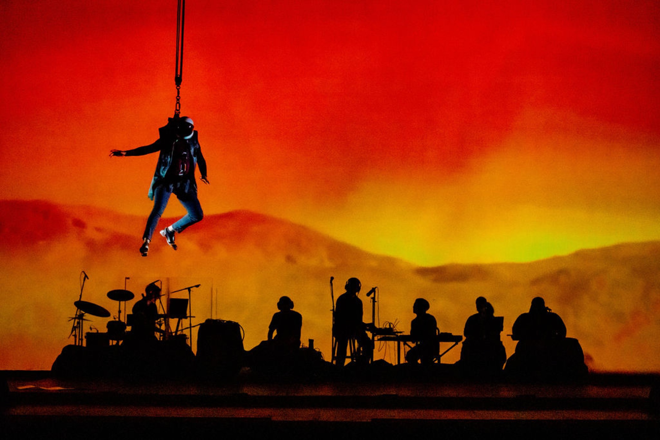 A performer suspended in a harness above silhouetted musicians in front of a red Mars-like backdrop