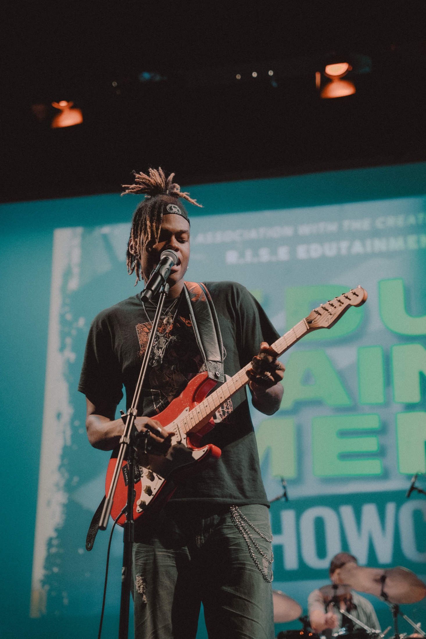 A guitarist singing into a microphone in front of a teal projected backdrop, drummer behind
