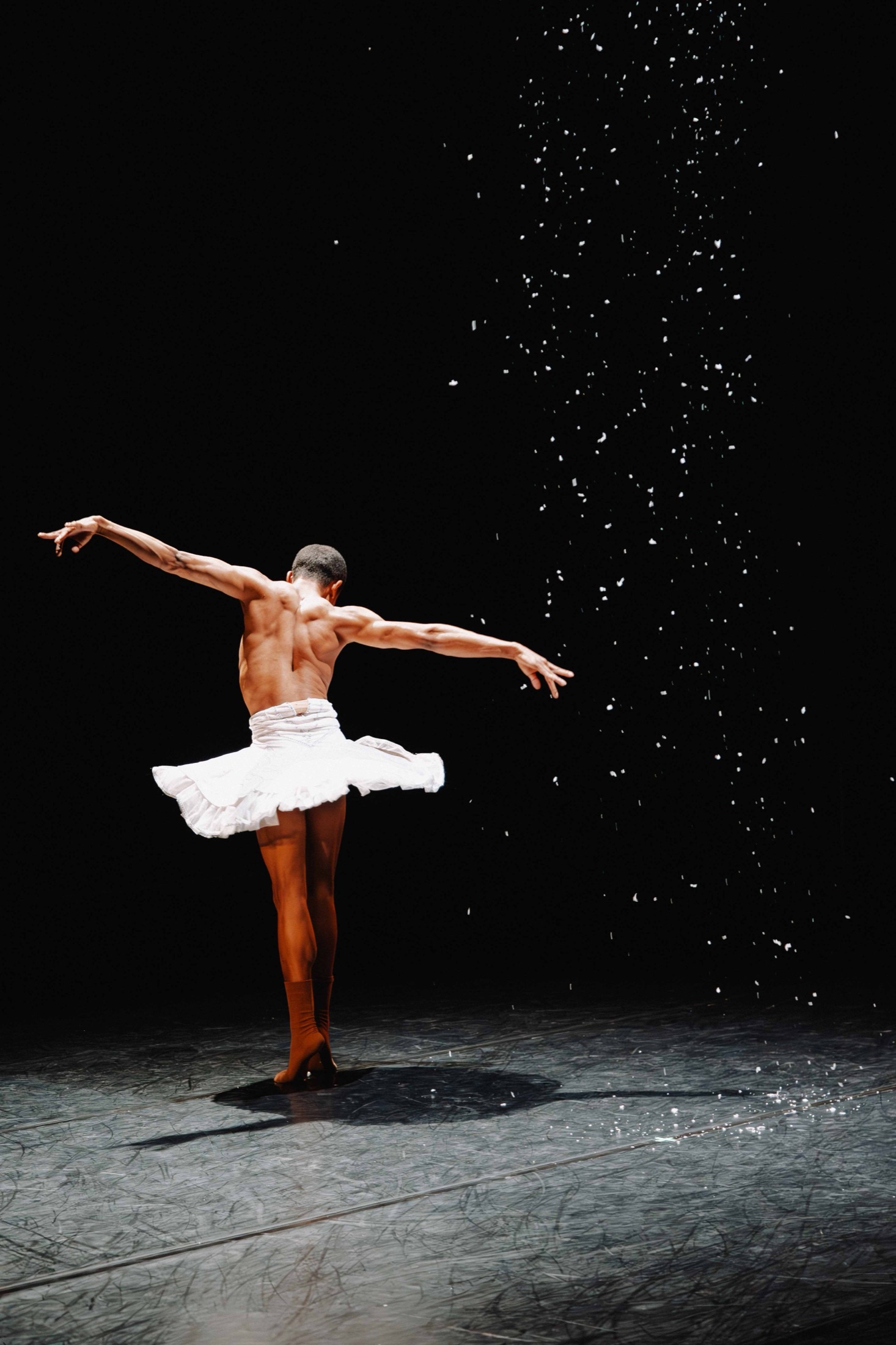 A male ballet dancer in a white tutu with his back to the camera under falling confetti