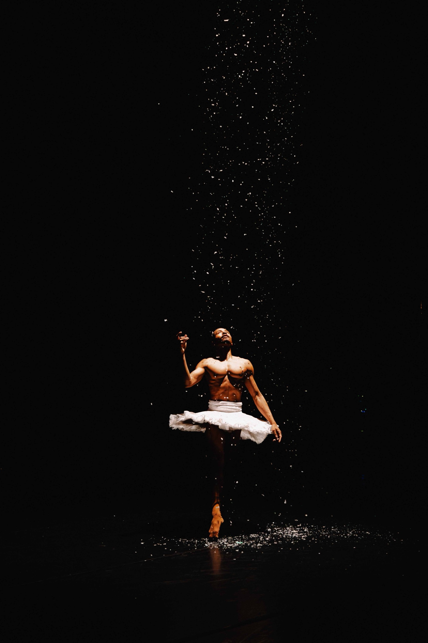 A male ballet dancer in a white tutu reaching upward as confetti falls around him